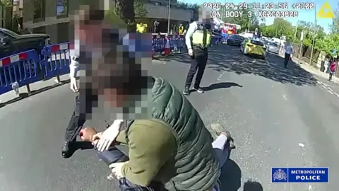 A man with a blurred face wrestles with a man on the ground as police officers stand nearby on a still from police bodycam footage