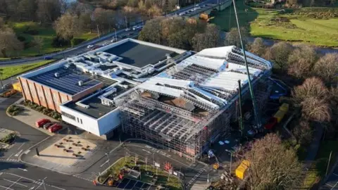 Aerial view of the Sands Centre taken in January after the storm. The right-hand side of the venue is covered with scaffolding, as is the section of roof above. Parts of a temporary canopy appear to have blown off. To the front and side of the building is a car park, while fields, a river and trees can be seen in the background.