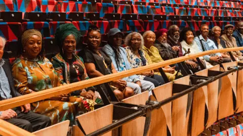 Bristol Beacon A long row of Bristol Boycott pioneers and family representatives sitting together in a theatre with further empty rows of blue and red geometric-patterned chairs behind them. Several of the men and women are wearing traditional Caribbean clothes which are brightly coloured