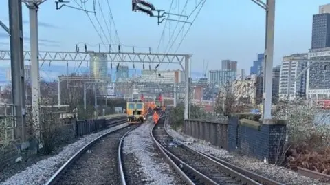 Two sets of rail tracks can be seen going into the distance, where a maintenance train and staff can be seen working. In the background is Birmingham's city skyline. 