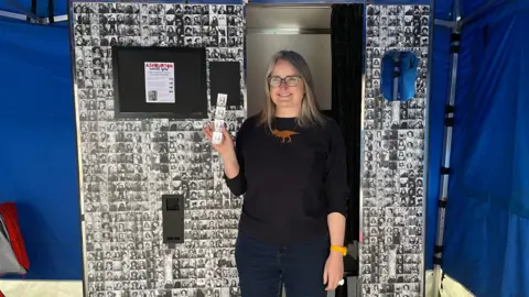 Artist Kate Tyler stading in front of a vintage photo booth, holding up a strip of four passport-style photographs. The photo booth is covered in similar photo strips.