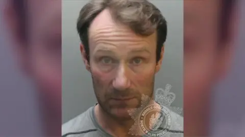 North Wales Police Christian Williams is a white man with short light brown hair. He is unshaven and wears a grey top. It is a head and shoulders police custody shot with a plain grey background and a watermark with the North Wales Police/Heddlu Gogledd Cymru crest on the lower right hand side of the photo.