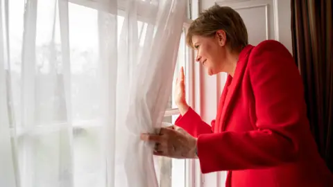 PA Media Nicola Sturgeon waves through a window at Bute House after announcing her resignation as first minister in February 2023. She is pulling aside the net curtains with her left hand and waving with her right hand.