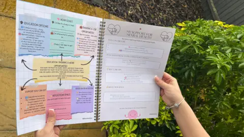 ELLEN KNIGHT/BBC A photo taken over the shoulder of Shannon, who's holding open the booklet. On one page are brightly coloured boxes that prompt users to write in their career goals, and explain different education options. On the other page, resources for support with mental health are written out. 