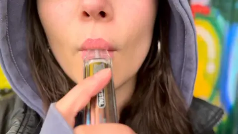 An anonymous teenage girl holds a vape to her lips, just the lower half of her face visible. She is in a purple hoodie, hood up, and black jacket and has long brown hair. She is in a park with colourful graffiti behind her.