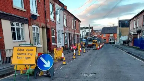 A narrow residential street which is partly taken up by road signs, cones and machinery