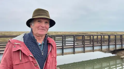 Edd Smith/BBC Ian Curtis wearing a khaki-coloured hat, a blue jumper and red raincoat. He is standing in front of the new Stiffkey bridge.