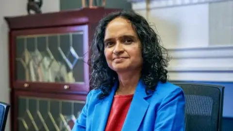 MacArthur Foundation Shailaja Paik seen in an office wearing a red dress and a blue blazer