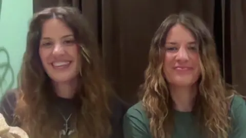 Georgia and Melissa Laurie, both sat in front of a webcam, smiling for the camera as they speak during an interview with BBC Breakfast. They both have long, brown or brown/blonde hair. Georgia is wearing a black T-shirt and Melissa a green T-shirt.