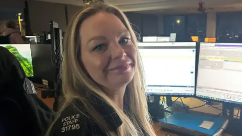 A woman with blonde hair sitting in front of a computer in a police control room