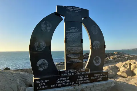 Johannes Dell/BBC A large, horseshoe shaped marble stone commemorating the 1904–1908 genocide in Namibia, stands on barren rocks against the backdrop of the ocean. The inscription reads: ‘Shark Island Luderitz. In memory of the Naman and Ovaherero Genocides. Here stood the Shark Island concentration camp, initially established in early 1905 as a concentration camp for the Ovaherero slave labourers for the construction of the Luderitz–Aus railway. … extermination meted out through slave labour, starvation, rape, exposure to adverse conditions and killing...