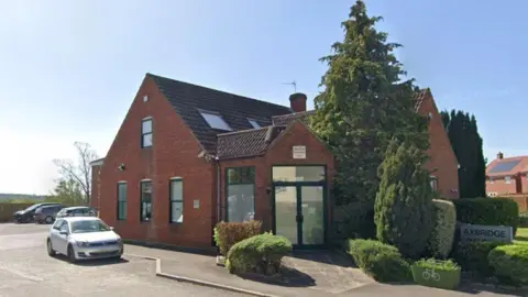 A redbrick building with brown-framed window panes. There is a large tree next to the entrance and a silver car is parked to the left of the building. The picture was taken on a sunny day.