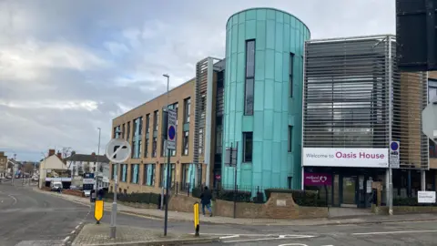 Pete Cooper/BBC A brick building with a turquoise blue cylinder on the side. The building has a large sign reading: "Welcome to Oasis House". There is a person walking past the building on the pavement. 