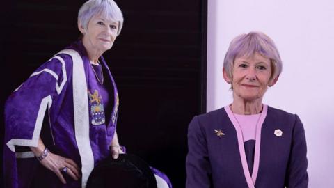 Prof Dame Nancy Rothwell, smiling with grey hair and wearing a pink-trimmed purple jacket and skirt, stand in front of a portrait of herself wearing her purple and silver ceremonial robes