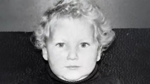 A black and white image of a young boy with blond hair, looking into the  camera with a smile