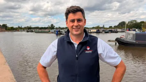 A man wearing a white shirt and a navy blue gilet is standing by a body of water with boats moored behind him. He has his hands on his hips and is smiling for the photograph.