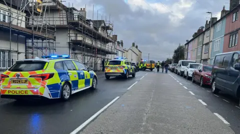 London Road in Chippenham with houses on either side and cars parked. There are three police cars with sirens stopped on the road.