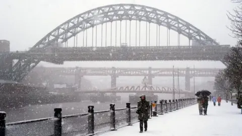 Snow covers the Newcastle Quayside. People walk by the river wearing thick coats and holding umbrellas. The Tyne Bridge, Swing Bridge and High Level Bridge can be seen in the background.