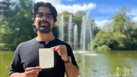 A man in a black top holds up a piece of paper with typewritten script. Behind him is a lake with jets spouting water. The lake is surrounded by mature trees.