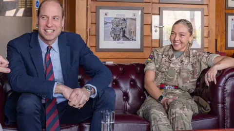 Prince William sits on a burgundy, leather Chesterfield sofa in an army barracks, talking to someone sitting across from him. He is wearing a navy suit, light blue shirt and a navy and red tie. Sitting beside him is a girl in her early 20s who is wearing a camouflage army uniform with her blonde hair slicked back into a ponytail. She is smiling with her arm resting on the arm of the sofa. There are framed army photographs hanging on the wood panelled wall behind them.