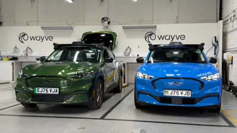 A shiny green car is in a garage next to a shiny blue car. There are small cameras on the roofs of both. 