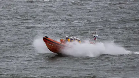 RNLI/Dave Cocks The Atlantic 85 Leicester Challenge III, an orange boat, on the water. White spray is around the boat. Four RNLI volunteers can be seen on the boat with yellow helmets and yellow jackets on.