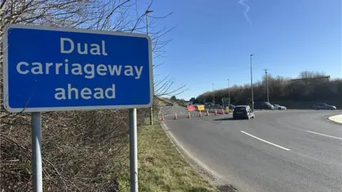 The eastbound carriageway of the A38 is closed following a crash near the Twelvewoods roundabout in Dobwalls, Cornwall. A line of traffic cones have been placed along the road of one of the roundabout exits. Signs saying the road is closed and a diversion has been set up have been erected. A blue highways sign saying "Dual carriageway ahead" is in the foreground. Several vehicles are entering the roundabout and a black Ford car is driving around the bend.