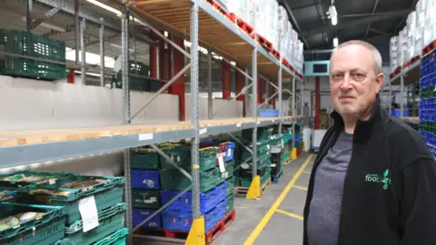 A man with grey hair and a black jacket stands in a warehouse. Rows of empty shelves and stacks of crates can be seen behind him.