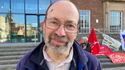 Jon Dale from the Unite union is wearing glasses and a beard. He is taking part in a protest outside the Derby City Council building. 