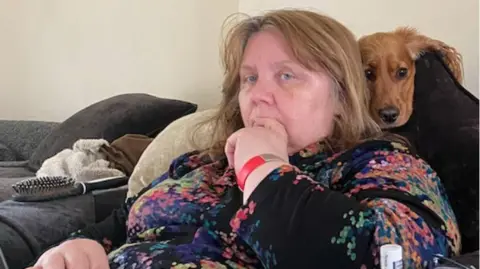 Beverley Cuddy A woman sat on a sofa with her finger in her mouth and a dog resting behind her head.