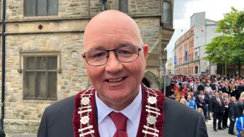 William Walker is smiling and wearing glasses. He is wearing a red collarette and a grey suit with a red tie.