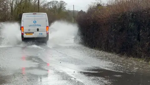 A white van drives through surface water on a rural road lined by hedges. The water is splashing up both sides of the vehicle.