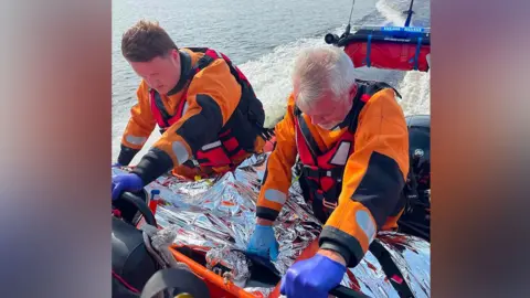 Two men wearing orange hi-vis jumpsuits and life jackets on board a rescue boat. They are treating a person on a stretcher who is wrapped in a foil thermal blanket - their face is not visible