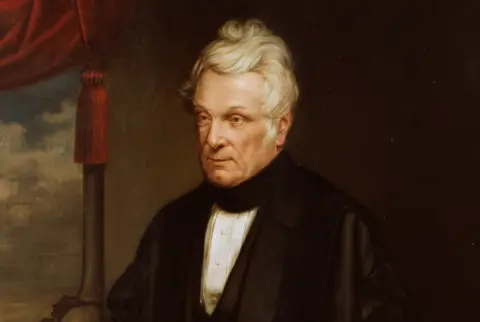English Heritage A painted portrait of John Clayton who has white, sweeping short hair. He is wearing a black coat and scarf with a white shirt underneath. He is sitting in front of a painted background and a red curtain can be seen in the top left corner.