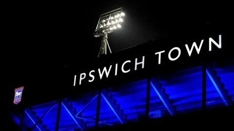 A view of Portman Road stadium at night. The club's name, Ipswich Town, is lit up in big white letters. The club logo sits to the left of the words on the stand. A flood stand with flood lights sit above.