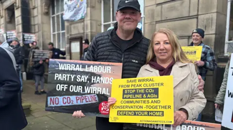 Gill Antrobus has long blonde hair and is wearing a beige trench coat . Her partner Scott Ball is wearing a black coat and baseball cap. Both are holding up signs protesting against the Peak Cluster project. They are among protesters outside Wallasey Town Hall. 