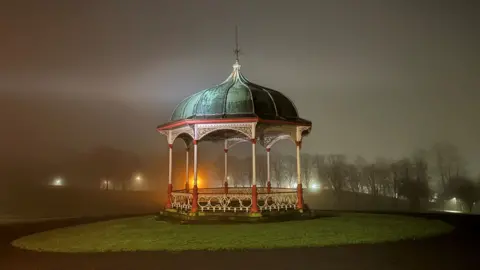 A Victorian-style bandstand stands illuminated in a misty park at night. 