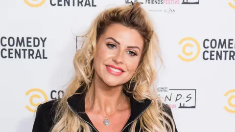 A blonde woman with a half up half down pony tail. She is wearing a silver necklace and a black jacket. She is smiling at the camera 