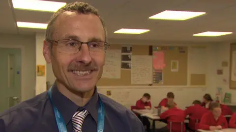 A male teacher with short brown hair and a moustache smiles as he stands in a classroom at his primary school. He is wearing a blue shirt and white and white tie with blue and brown stripes. In the background, children wearing red uniforms sit working around tables.