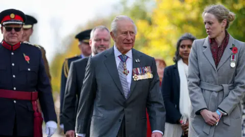 PA Media King Charles III arrives for a visit to the National Memorial Arboretum in Alrewas, Staffordshire, for the dedication ceremony of the LGBT+ Armed Forces memorial, the UK's first national memorial commemorating LGBT+ people who have served and continue to serve in the military. He is seen walking through the arboretum wearing a dark suit over a white shirt, wearing his military medals.