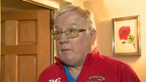 A man stands in the hallway of his house. He has short white hair and wears clear glasses. Over his left shoulder there is a portrait of a red poppy with a green stem. Over his right shoulder there is an ajar brown wooden door.