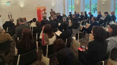 Government of Jersey A large group of people site inside a room with white walls while two artists perform in front of them.