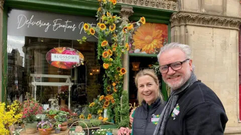 A man wearing glasses with his arm around a woman standing outside a florist's shop with various floral displays on the pavement