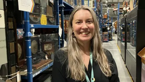 A woman looks into the camera, smiling. She is standing amongst exhibits at the National Railway Museum, with a model of the London Victoria Line and clocks behind her. Next to those is a white board in the shape of an arrow, with rainbow trim.