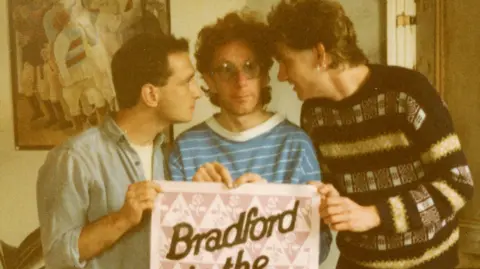 Alan Richardson/Castles in the Sky Three men stand together holding a pink and white poster. It is a sepia-toned image from the 1980s