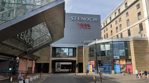 Google Maps A large shopping centre building, with ST ENOCH and VUE written in large letters on one wall. A road runs along the side of the building and passers-by are walking along the street nearby. 