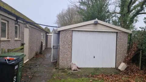 LDRS A garage in pale pebble-dash with a white door is on the right of the image. To the left is the side wall, also pebble-dashed, of a church hall.