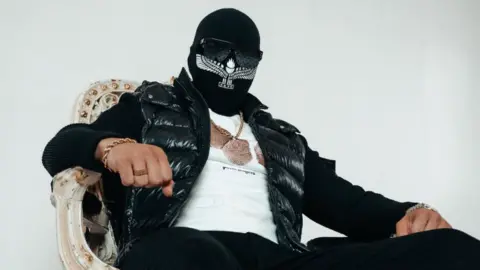 Gaboro, sat on a chair, wearing a balaclava, sunglasses and a chain