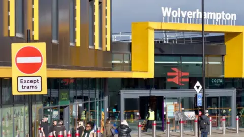 Wolverhampton train station has yellow/gold rail along the front and raised above the glass entrance. There are people entering and leaving the station via the glass doors. The word Wolverhampton is visible above the raised yellow rail and is in white writing.