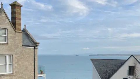The view from the garden at Talland House with Godrevy lighthouse in the distance across the sea - all seen in the gap between two buildings, one of which has a balcony with potted plant on it.